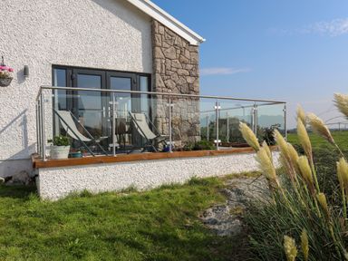 A balcony with two lounge chairs and glass railing outside a house with stone and white walls at Corrie Knowe in Trearddur Bay
