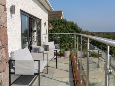 An outdoor patio with grey cushioned seats and glass railing overlooking greenery at Corrie Knowe in Trearddur Bay