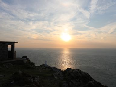 A coastal scene with a stone structure a lighthouse on a cliff and the sun setting over the ocean at Corrie Knowe in Trearddur Bay