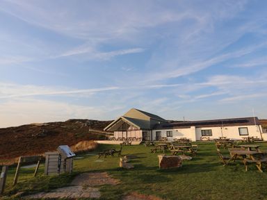 An outdoor seating area with wooden tables and chairs on grass near a white building with a slanted roof in a rural setting