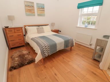 A bedroom with a wooden double bed, two bedside tables with lamps, a chest of drawers, a window with a teal blind, and a rug on a wooden floor at Little Tern in Seahouses