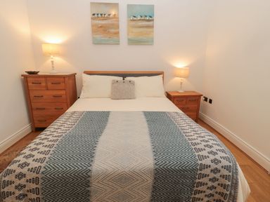 A bedroom with a bed between a chest of drawers and a bedside table with lamps and paintings on the wall at Little Tern in Seahouses