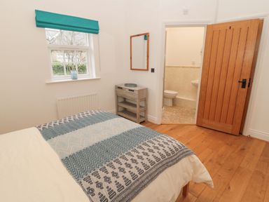 A bedroom with a bed covered with a patterned blanket a small wooden cabinet a wall mirror an open wooden door leading to a bathroom and a window with a teal blind at Little Tern in Seahouses