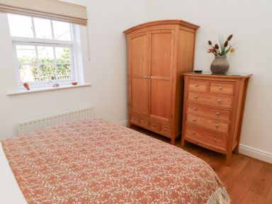 A bedroom with a window a bed with a patterned cover a wooden wardrobe and a wooden chest of drawers with a vase with flowers on top at Little Tern in Seahouses