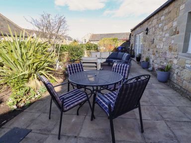 An outdoor patio with a round metal table and four cushioned chairs next to a stone building and garden at Little Tern in Seahouses