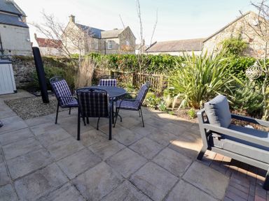 A patio area with a round table and four chairs next to a garden with plants and a cushioned bench at Little Tern in Seahouses