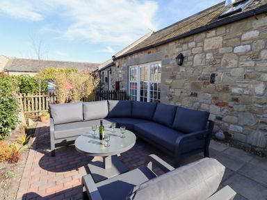 An outdoor seating area with a sectional sofa a round table and a chair on a brick patio by a stone building at Little Tern in Seahouses
