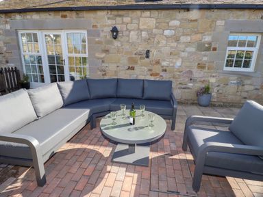 An outdoor seating area with grey cushioned sofas and a round table with a wine bottle and glasses on a patio at Little Tern in Seahouses