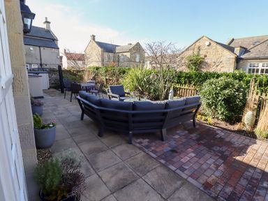 An outdoor patio area with a curved sectional sofa a table and chairs stone buildings and greenery at Little Tern in Seahouses