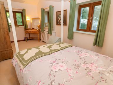 A bedroom with a bed covered in floral bedding and a green blanket green curtains wooden framed windows a wooden desk with a lamp and mirror a floral painting on the wall and a wooden wardrobe at Harveys House in Helston