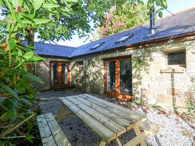 An outdoor patio area with a wooden picnic table and stone cottage walls at Harvey's House in Helston