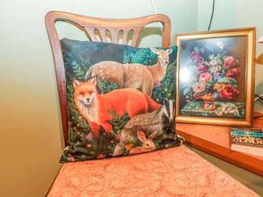 A wooden chair with a patterned cushion featuring animals and a wooden table with a framed floral painting and a book titled under dark skies at Harveys House in Helston