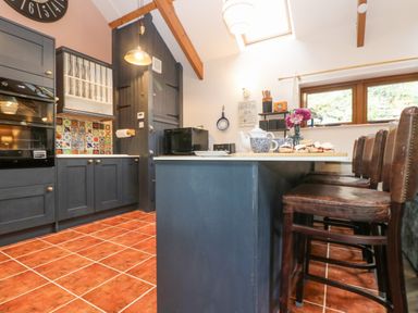 A kitchen with dark cabinets, a tiled floor, a kitchen island with a tea set and cakes, and bar stools at Harveys House in Helston