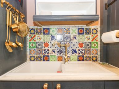 A kitchen sink with a brass faucet colorful patterned tile backsplash and utensils hanging on a rail at Harveys House in Helston
