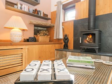 A living room with a lit wood stove next to a window and a tic tac toe game on a table at Harveys House in Helston
