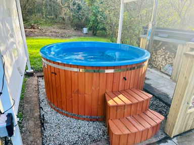 An outdoor hot tub with wooden exterior and steps surrounded by gravel and greenery at Harveys House in Helston
