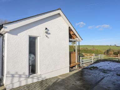 A white building exterior with a small porch and wooden railing next to a fenced area with a grassy field in the background at Capel Park (Tyddynod Bach) in Llandyfrydog near Llanerchymedd