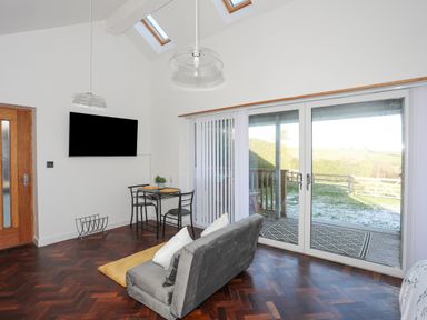 A living room with a gray sofa small dining table wall mounted tv and sliding glass doors at Capel Park Tyddynod Bach in Llandyfrydog near Llanerchymedd