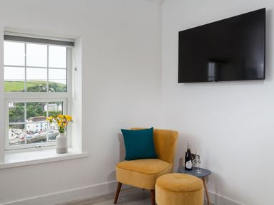 A corner with a yellow chair and ottoman a side table with a wine bottle and glass a window with flowers in a vase and a wall-mounted TV at Chy Lowen in Porthleven