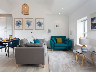 A living room with a grey sofa, a teal armchair, blue chairs around a dining table and framed artwork on the wall at Chy Lowen in Porthleven