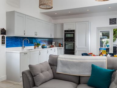 A kitchen with white cabinets a blue backsplash a gray sofa and a dining table visible through glass doors at Chy Lowen in Porthleven