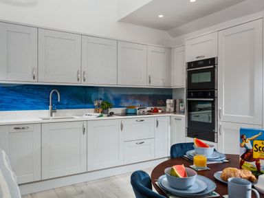 A kitchen with white cabinets a blue ocean-themed backsplash a round table with breakfast items including watermelon croissants and orange juice at Chy Lowen in Porthleven