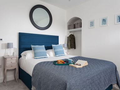 A bedroom with a blue upholstered bed and pillows a nightstand with a lamp an open closet with baskets and a jacket a round mirror and framed pictures on the wall at Chy Lowen in Porthleven