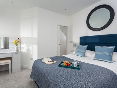 A bedroom with a bed a blue patterned blanket two pillows a round mirror and a white dressing table with flowers at Chy Lowen in Porthleven