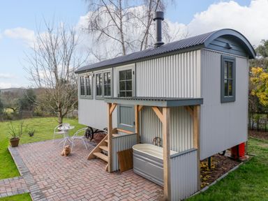 An outdoor patio with a small white table and chairs a wooden hut with corrugated metal sides and a covered bathtub on a paved area at Evendine Hut @ Evendine Cottage in Colwall Herefordshire