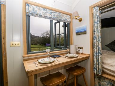 A small dining area with two stools and table set for two by a window with floral curtains at Evendine Hut @ Evendine Cottage in Colwall Herefordshire