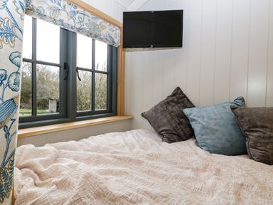 A bedroom corner with a bed covered in a beige quilt with pillows and a wall-mounted television near a window with floral curtains at Evendine Hut