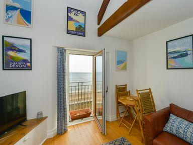 A room with a small dining table and chairs a leather sofa and an open door with a view of the sea at 2 Bayview in Torcross