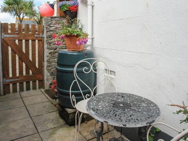 A small outdoor patio with a round metal table and two white metal chairs next to a white wall and a wooden gate with plants and a large green water barrel