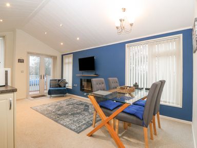 A dining area with a glass table and four chairs near a living space with a blue accent wall and a mounted tv at Y Caban in Brynsiencyn