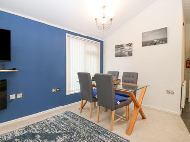 A dining room with a glass table and four grey chairs near a window with vertical blinds at Y Caban in Brynsiencyn