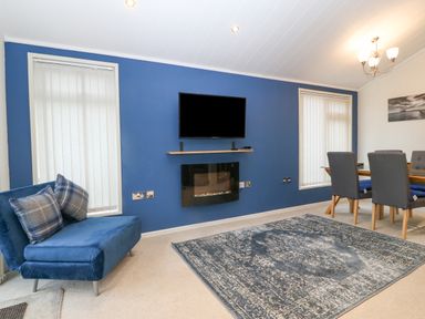 A living room with a blue accent wall a mounted television a fireplace a blue armchair with plaid pillows a patterned rug and a dining table with four chairs at Y Caban in Brynsiencyn
