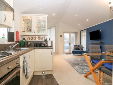 An open kitchen and living area with white cabinets glassware a blue sofa a wall-mounted television and a glass dining table at Y Caban in Brynsiencyn