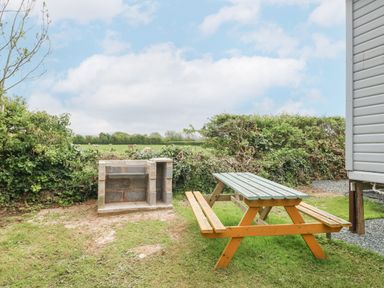 An outdoor area with a wooden picnic table and a brick barbecue grill surrounded by greenery at Y Caban in Brynsiencyn