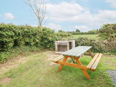 A picnic bench and a stone barbecue grill in a grassy outdoor area with hedges at Y Caban in Brynsiencyn