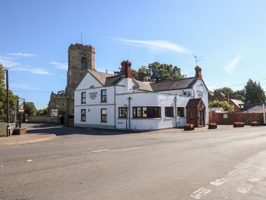 A white building with a sign reading Five Bells and an old church tower behind it at The Five Bells Inn in Upwell