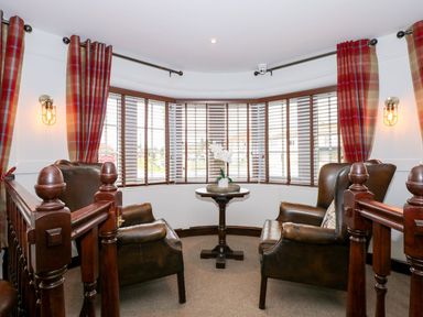A sitting area with two leather armchairs and a round table with flowers by a bay window with wooden blinds and red plaid curtains at The Five Bells Inn in Upwell