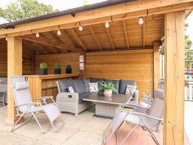 An outdoor seating area with a wooden roof and pillars a cushioned corner sofa a table and reclining chairs at The Five Bells Inn in Upwell
