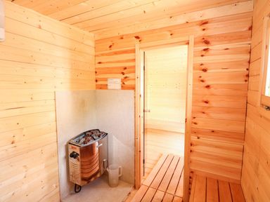 A wooden sauna room with a heater, stones on top, a plastic water jug, and a door leading to another wooden sauna room at The Five Bells Inn in Upwell