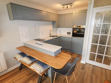 A kitchen with grey cabinets a wooden dining table with cushions on benches and a grey chair at 9 Oakley Wharf in Porthmadog