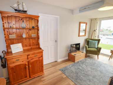 A living room with a wooden cabinet with model sailboats a green armchair with a pillow a small TV on a wooden chest and a sliding glass door overlooking water at 9 Oakley Wharf in Porthmadog