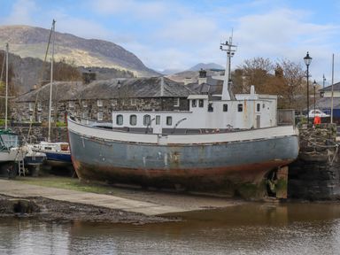 A boat on dry land near stone buildings and mountains in the background at 9 Oakley Wharf in Porthmadog