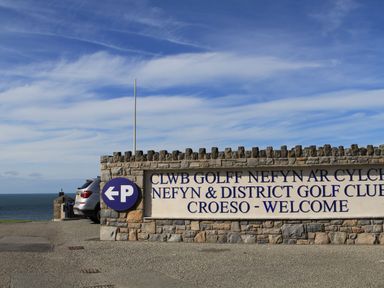 A stone wall sign for Nefyn and District Golf Club with parking sign and ocean view in the background