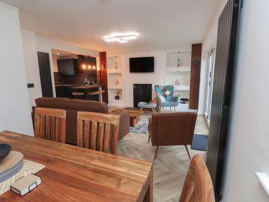 An open living room and kitchen area with wooden dining table wooden chairs a brown sofa brown armchair blue patchwork chair and a wall-mounted TV at Sea Cliff in Morfa Nefyn