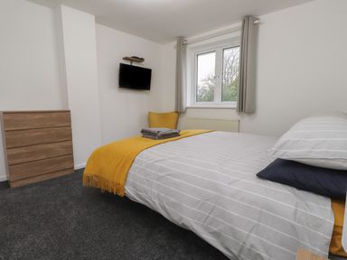 A bedroom with a bed covered in striped bedding and a yellow throw a wooden chest of drawers a wall-mounted tv and a window with grey curtains at Sea Cliff in Morfa Nefyn