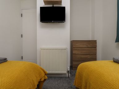 A bedroom with two beds covered in yellow blankets a wooden dresser a white radiator and a TV mounted on the wall at Sea Cliff in Morfa Nefyn